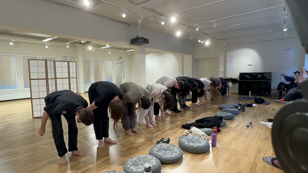 Actors bow in a curtain call line at the conclusion of 'Faith Hope Charity'. Cushions, water bottles, and other accessories are scattered on the floor in front of them. Audience members are pictured to the right of the performers, applauding.